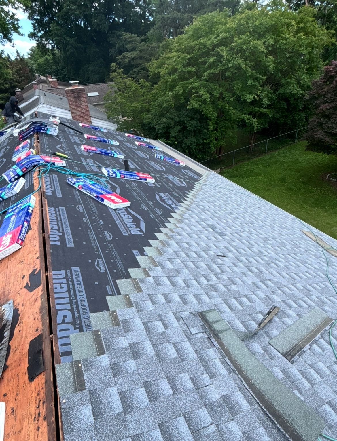 Roofing work in progress; new shingles on the right, underlayment on the left, trees in the background.