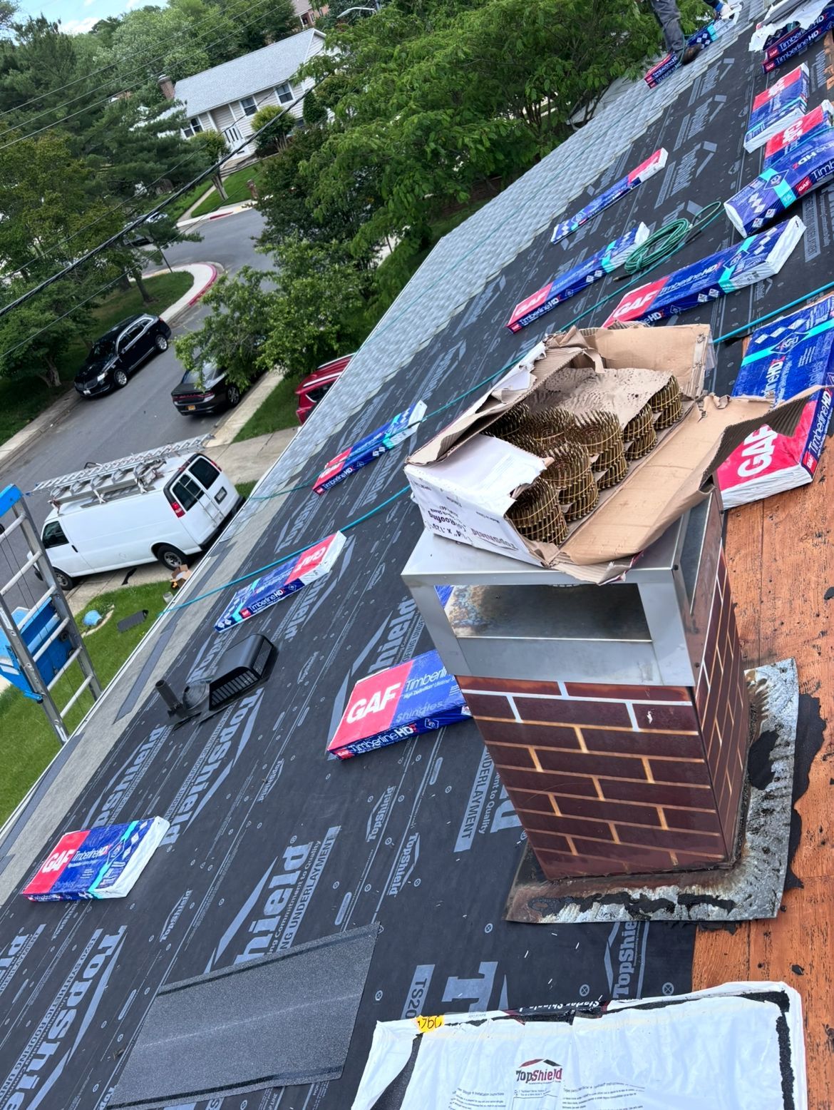 Roof with shingles being installed; chimney and cardboard box on top. White van and ladder on the side.
