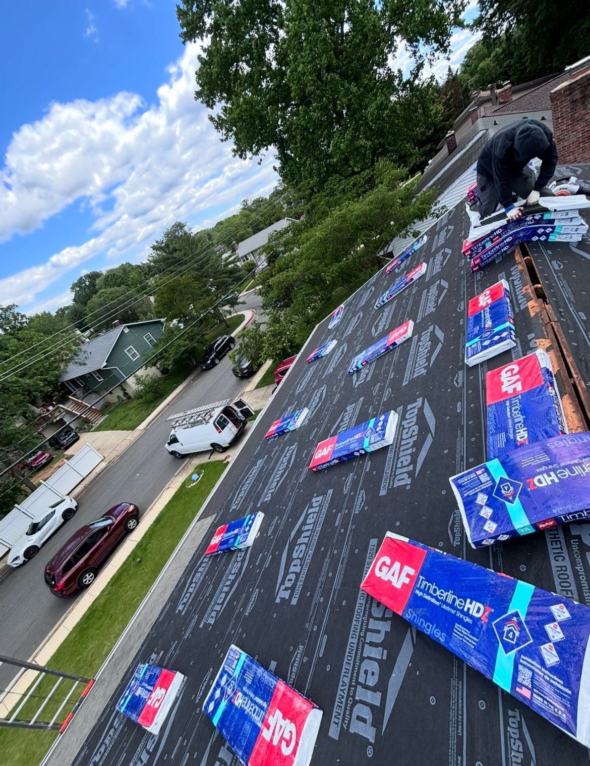 Roofer on a roof covered in GAF shingles, bags of shingles scattered. Street with parked cars in the background.