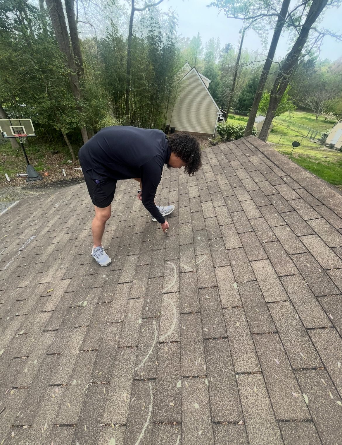 Person on a rooftop, inspecting shingles. Outdoor setting, trees in background.