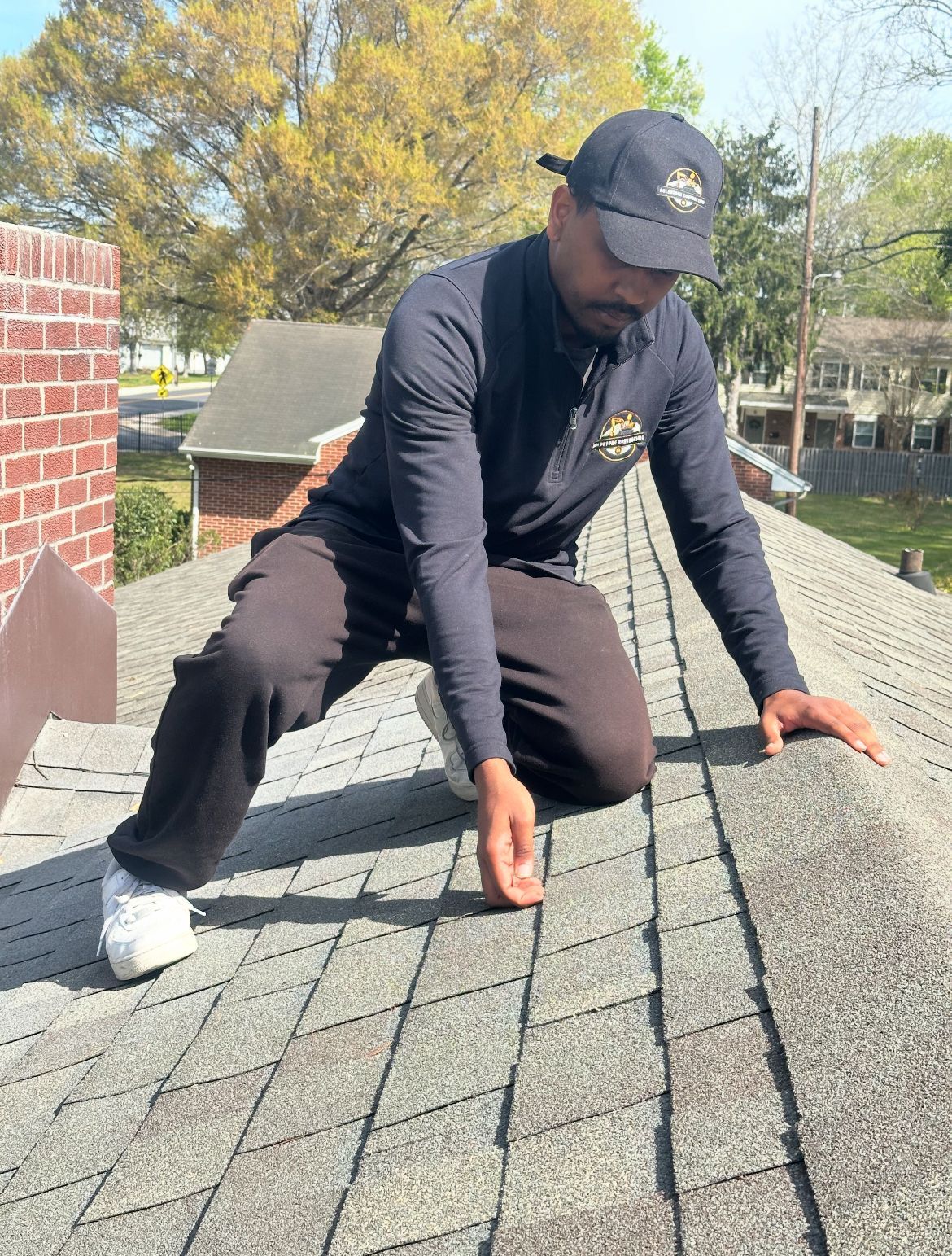 Person in cap and dark clothing inspecting asphalt shingle roof. Outdoors, sunny day.
