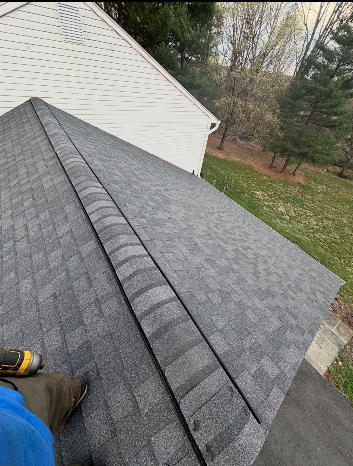 A person standing on a gray shingle roof. The roof has a ridge cap, and a white house and trees are visible in the background.