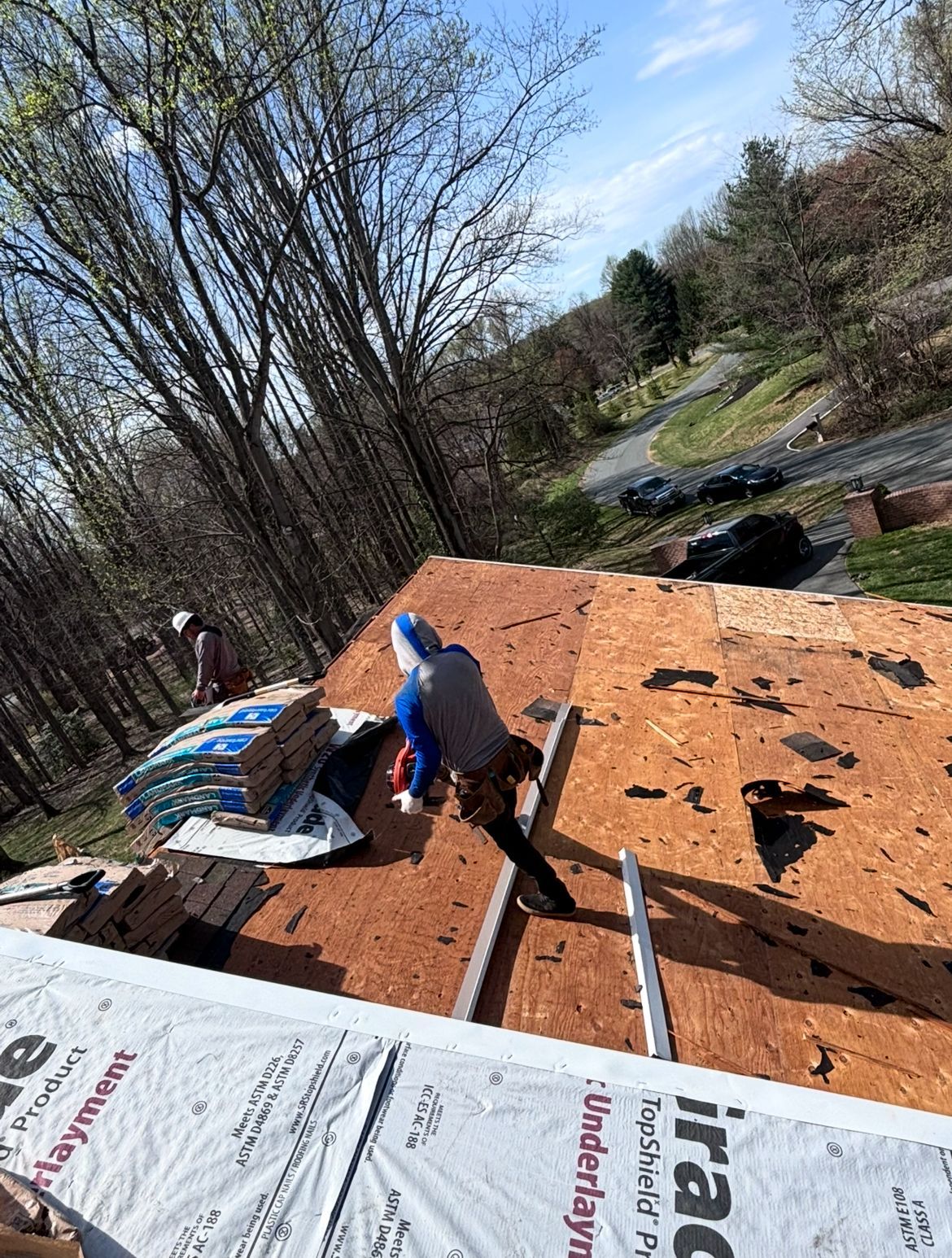 Roofers working on a house roof, holding materials, with a forested hillside in the background.