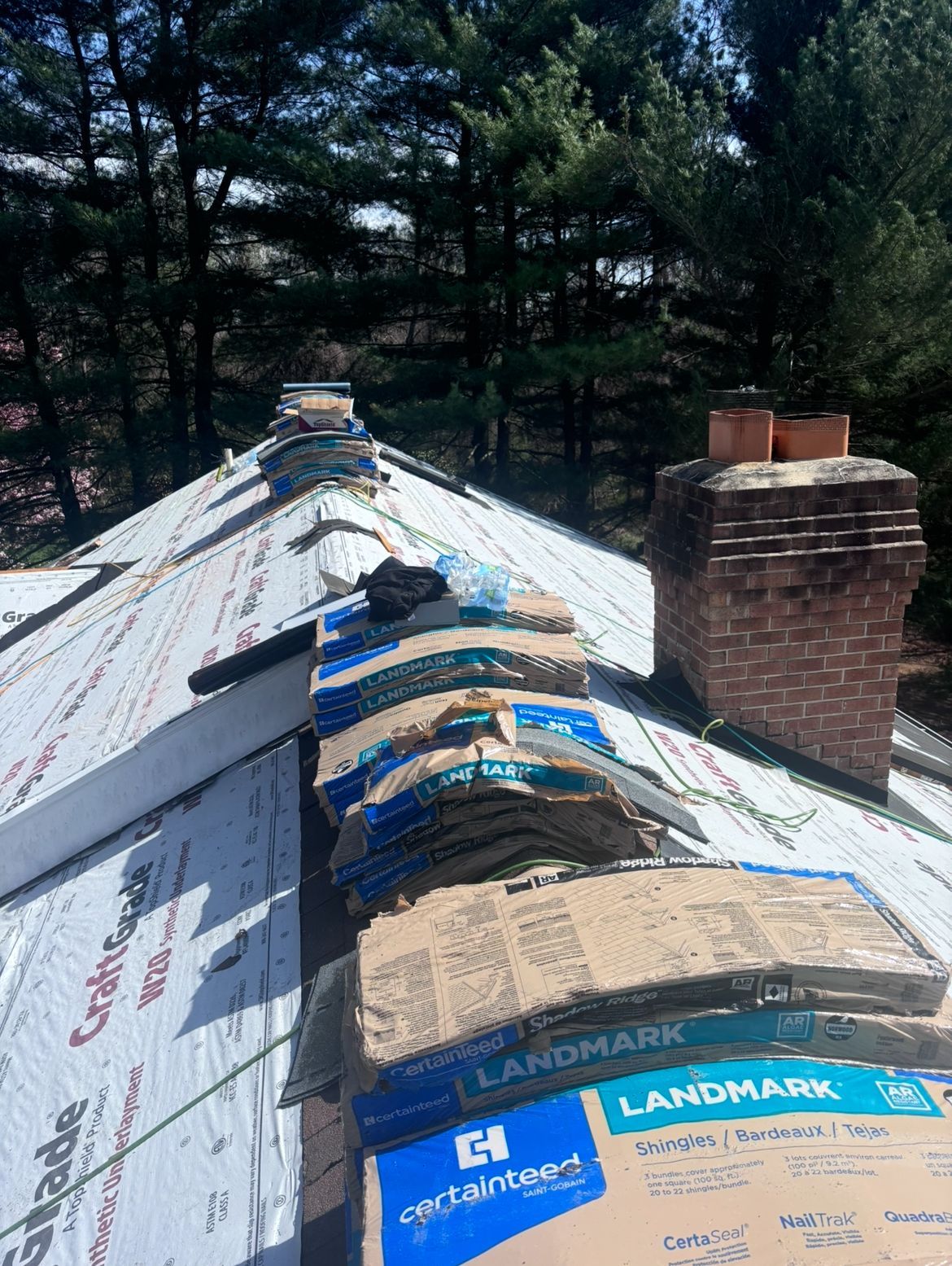 Rooftop with cardboard boxes of roofing shingles, near a chimney and trees.