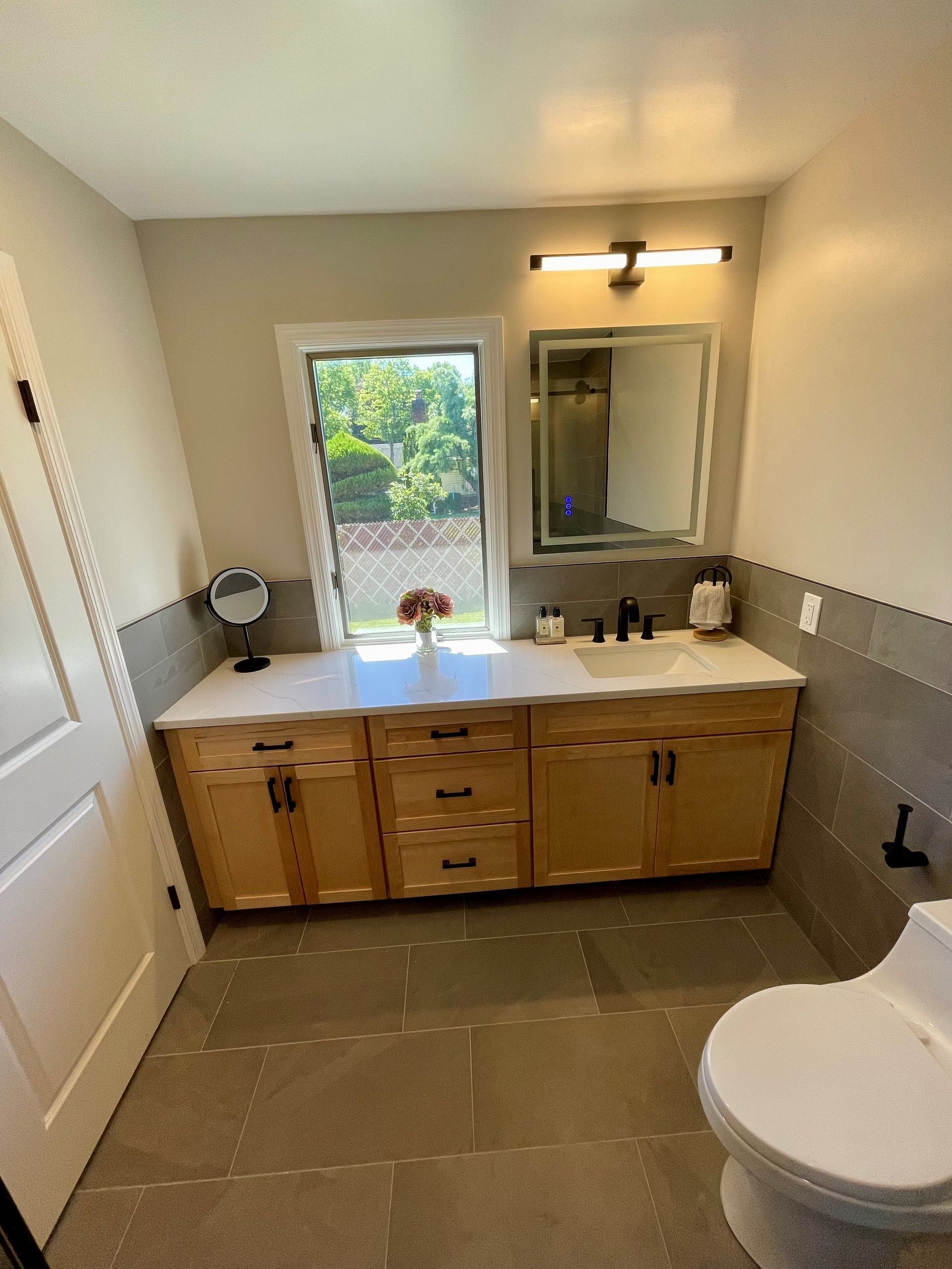 A bathroom with a toilet, sink, mirror and window.