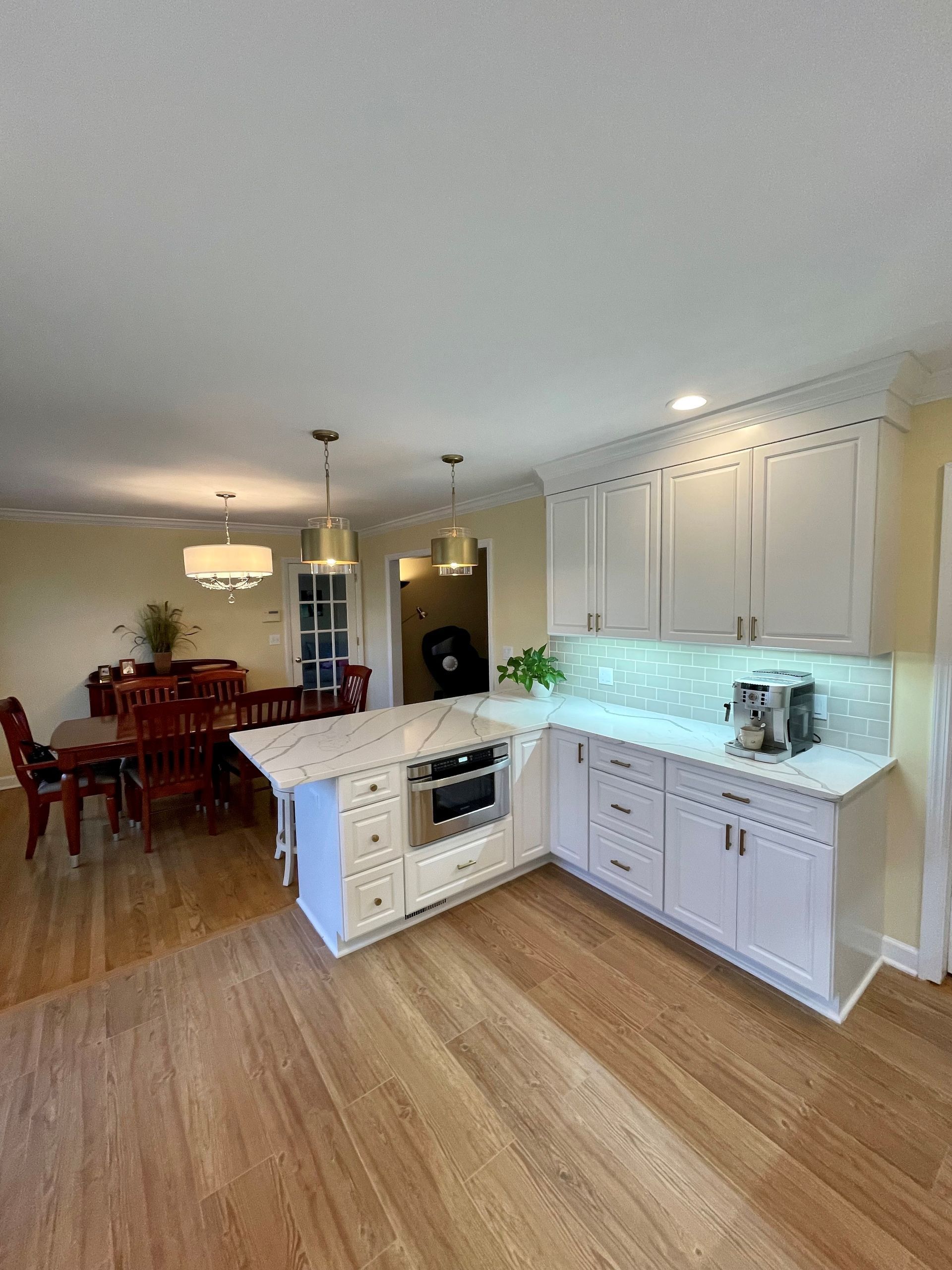A kitchen with white cabinets and hardwood floors and a dining room in the background.