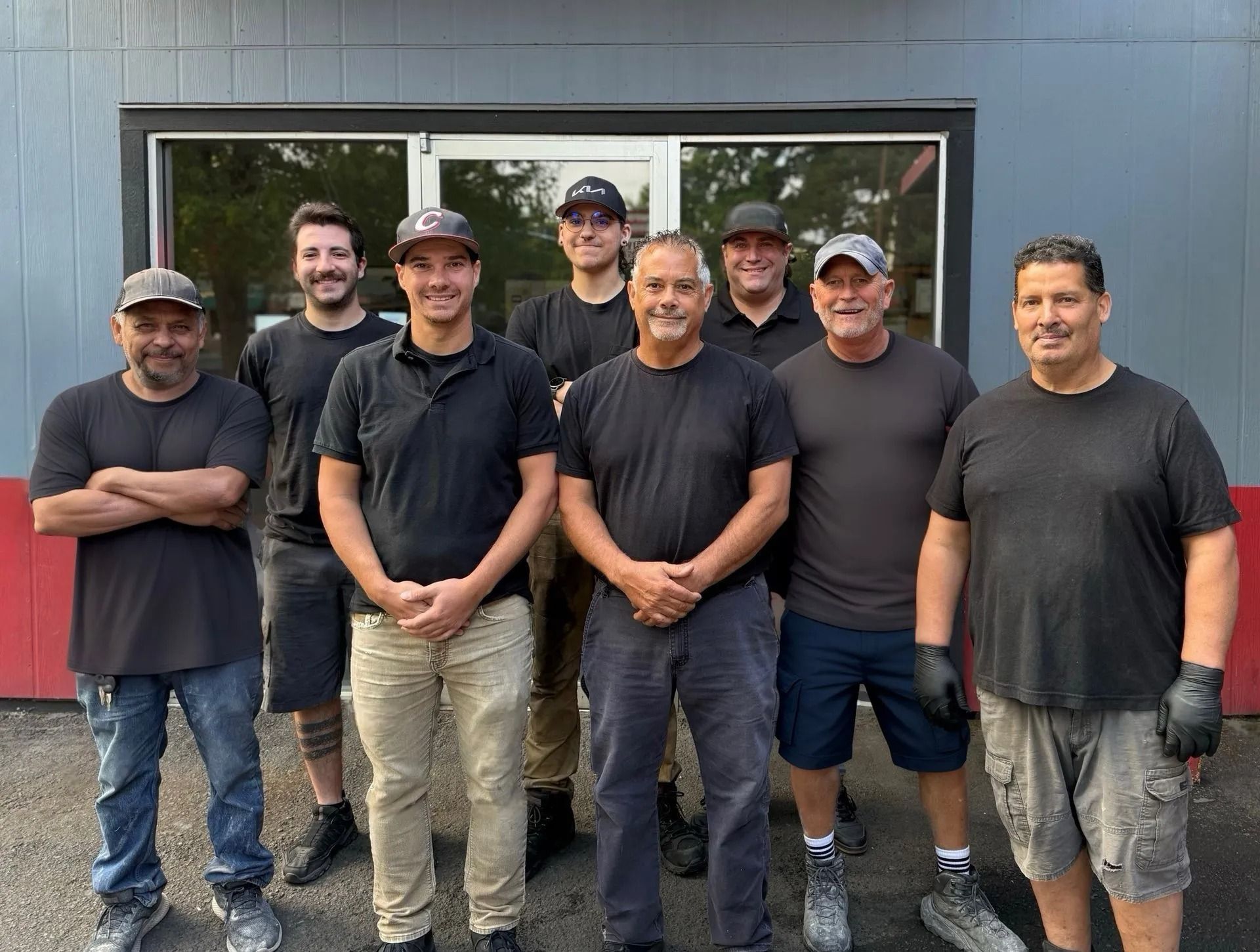 Group of men posing in front of a building. Most are wearing black shirts. Some are wearing hats.
