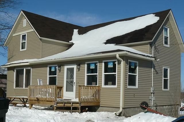 Beige house with porch, snow on roof and ground, blue sky.