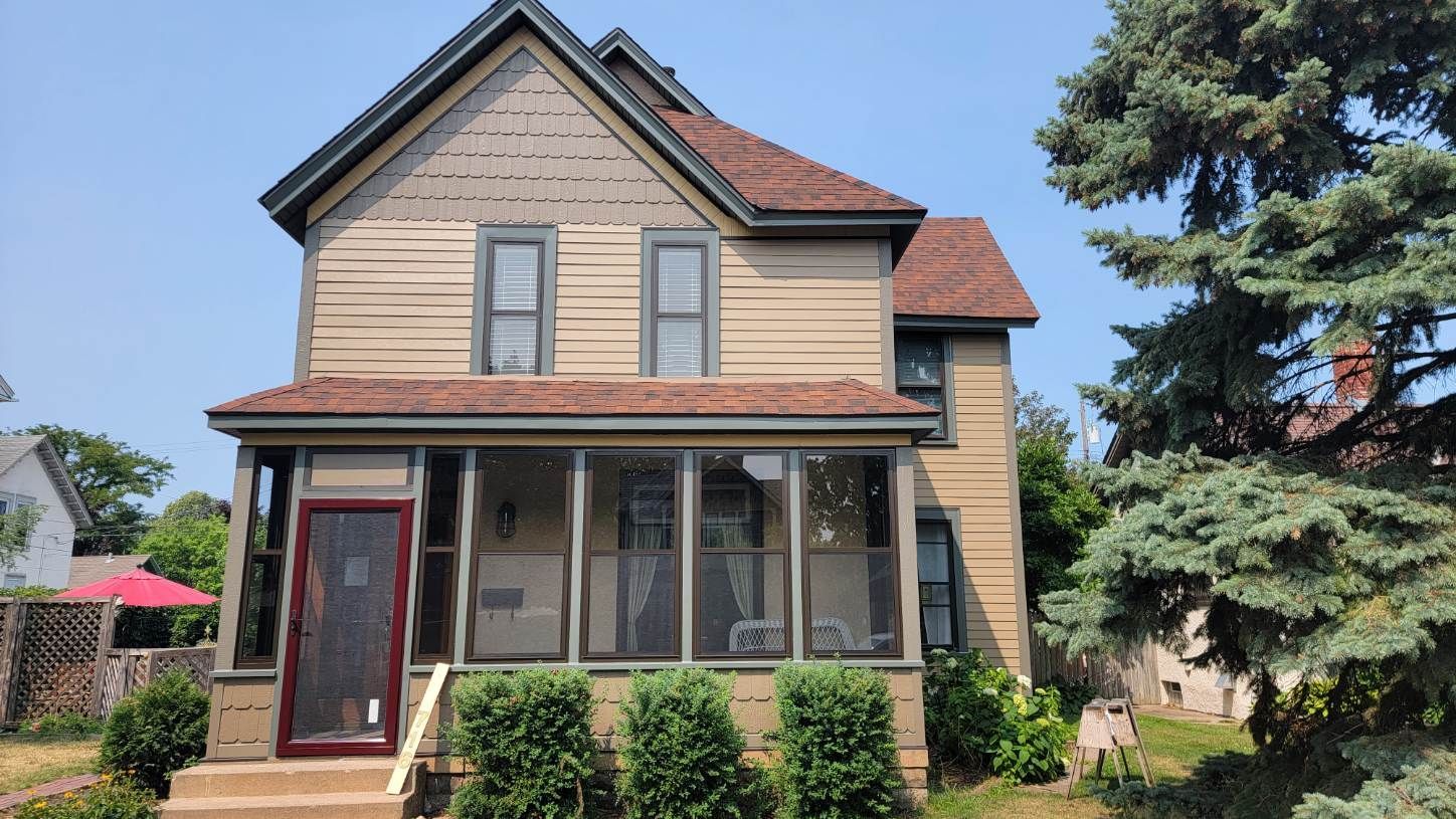 Two-story house with screened porch, brown roof, and tan siding. Green bushes in front and blue sky.