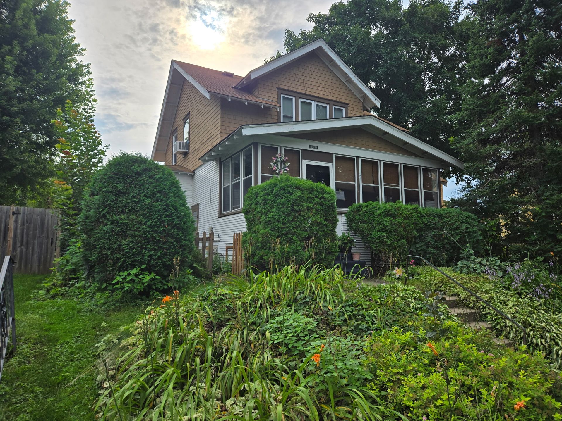 Two-story house with overgrown bushes and plants in front. Overcast sky.