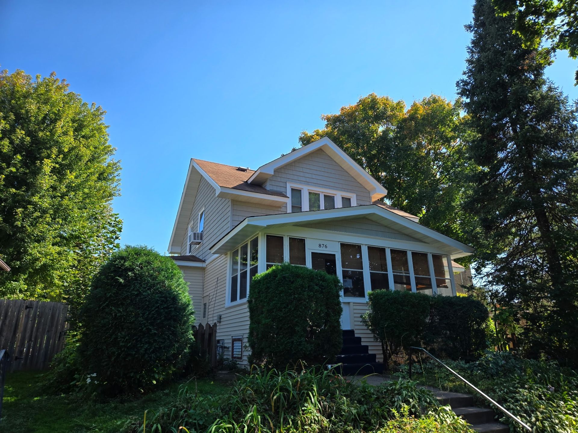 Beige two-story house with porch and bushes in front, trees on either side, under blue sky.