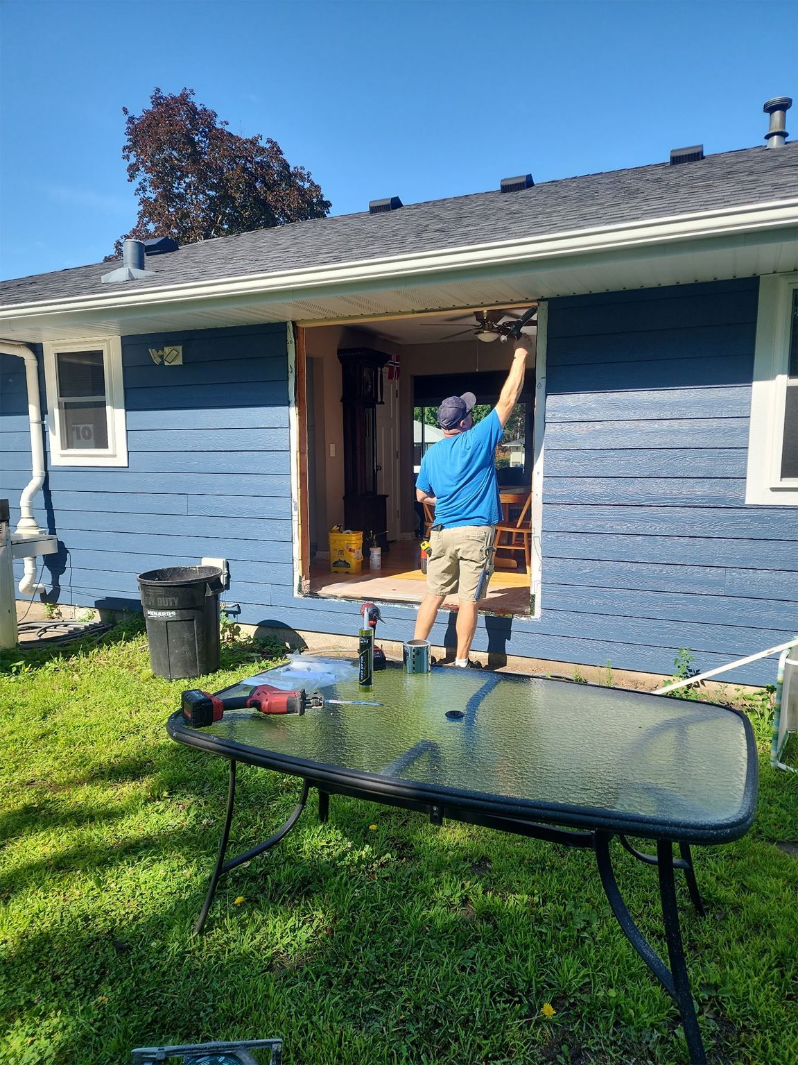 Person working on a doorway of a blue house; tools on a table in the foreground; sunny outdoor setting.