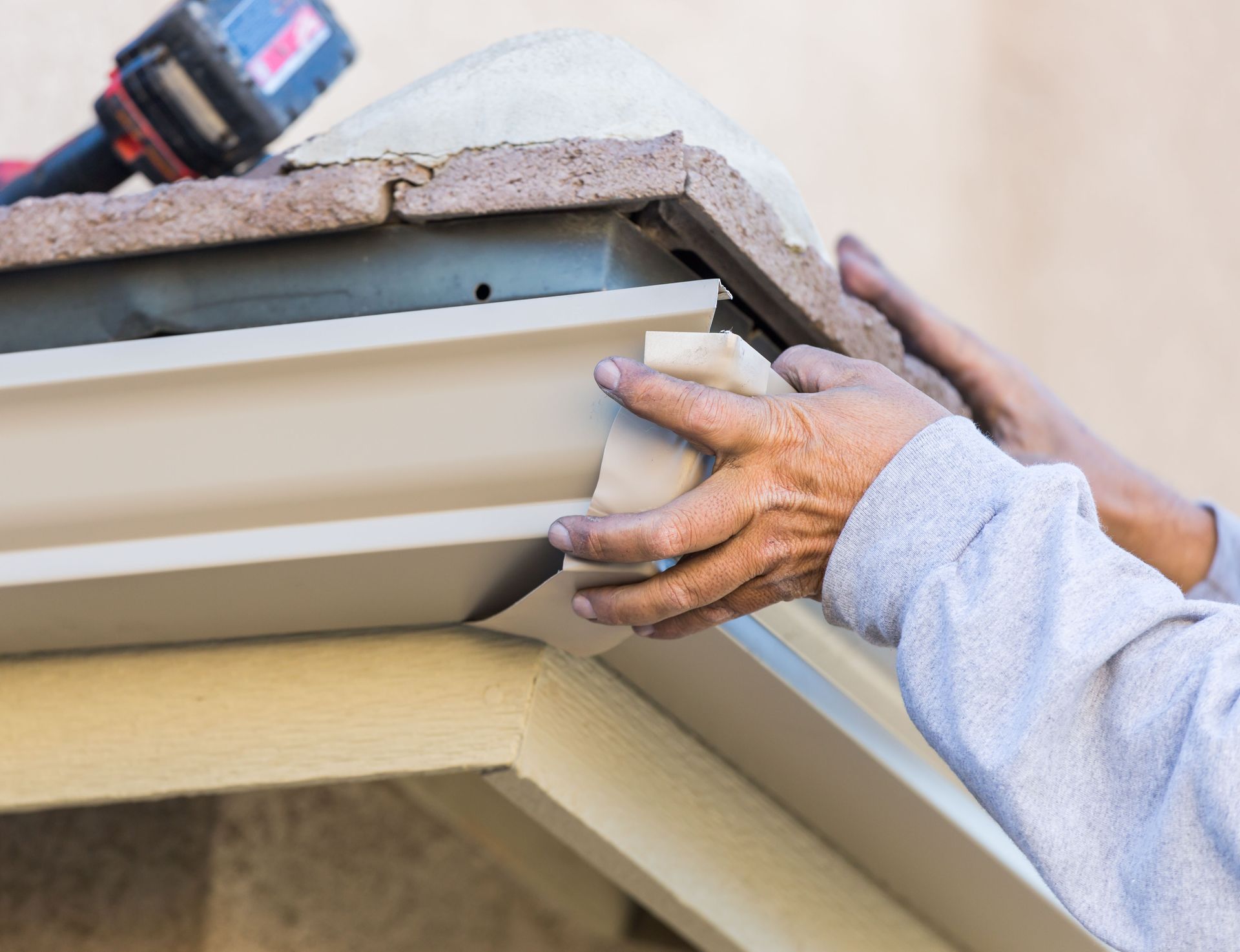 Hands installing beige gutter on a building's roof edge, with a power drill visible.