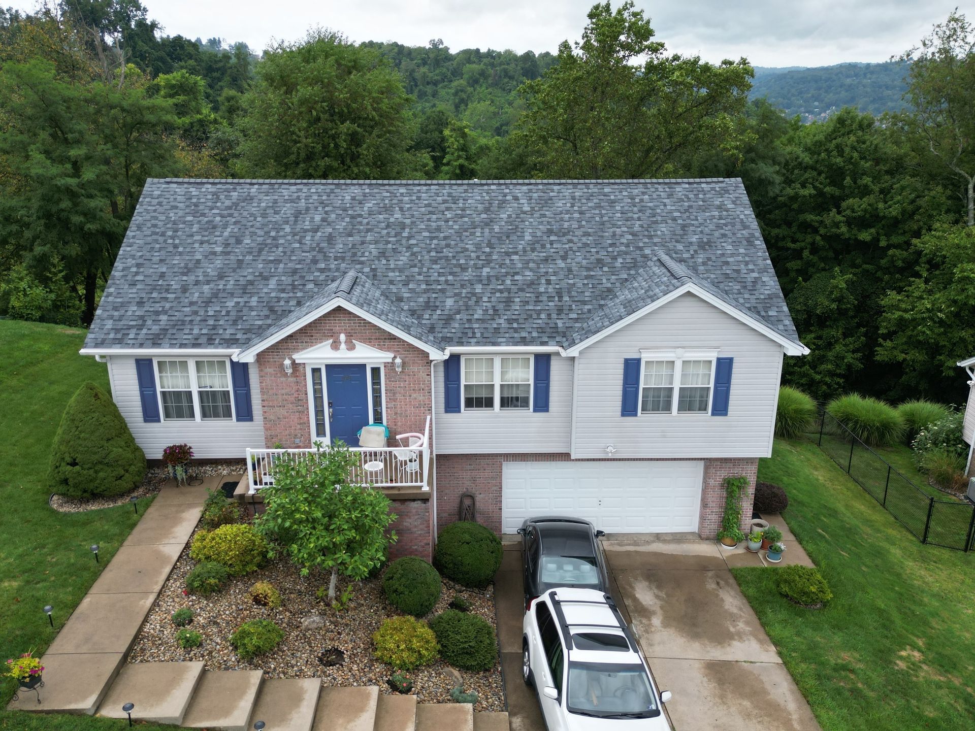 A house with a blue door, brick facade, and gray roof. Cars sit in the driveway. Green trees in the background.