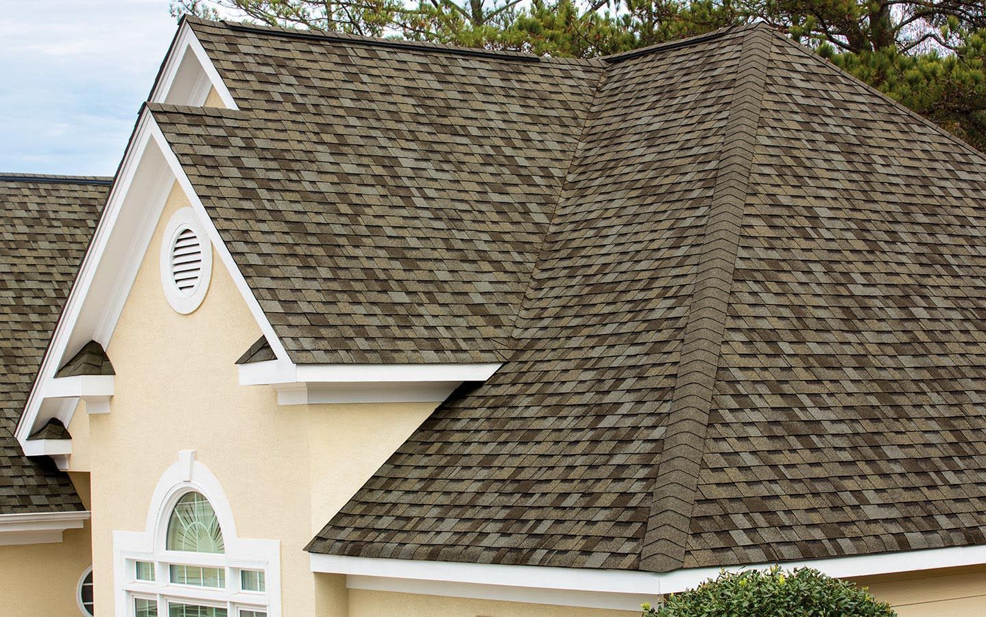Tan house with a weathered, brown shingle roof, white trim, and a cloudy sky.
