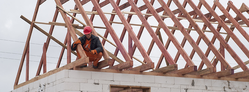 A construction worker wearing orange overalls and a red hat on a partially built roof frame.