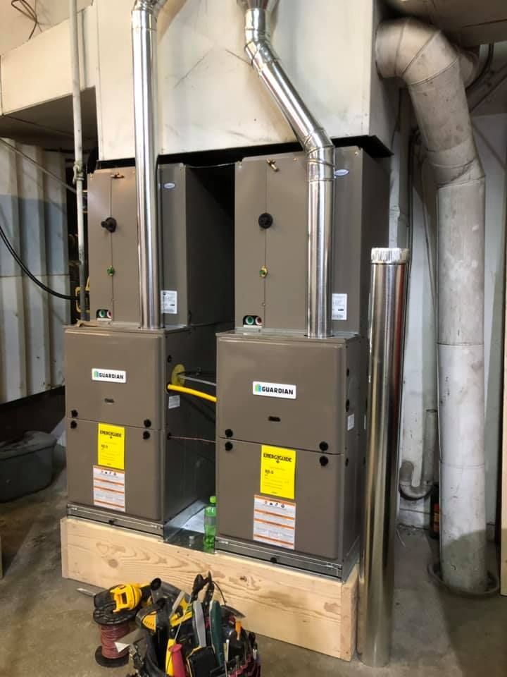 Two gray furnace units in a utility room with exposed ductwork and tools on the floor.