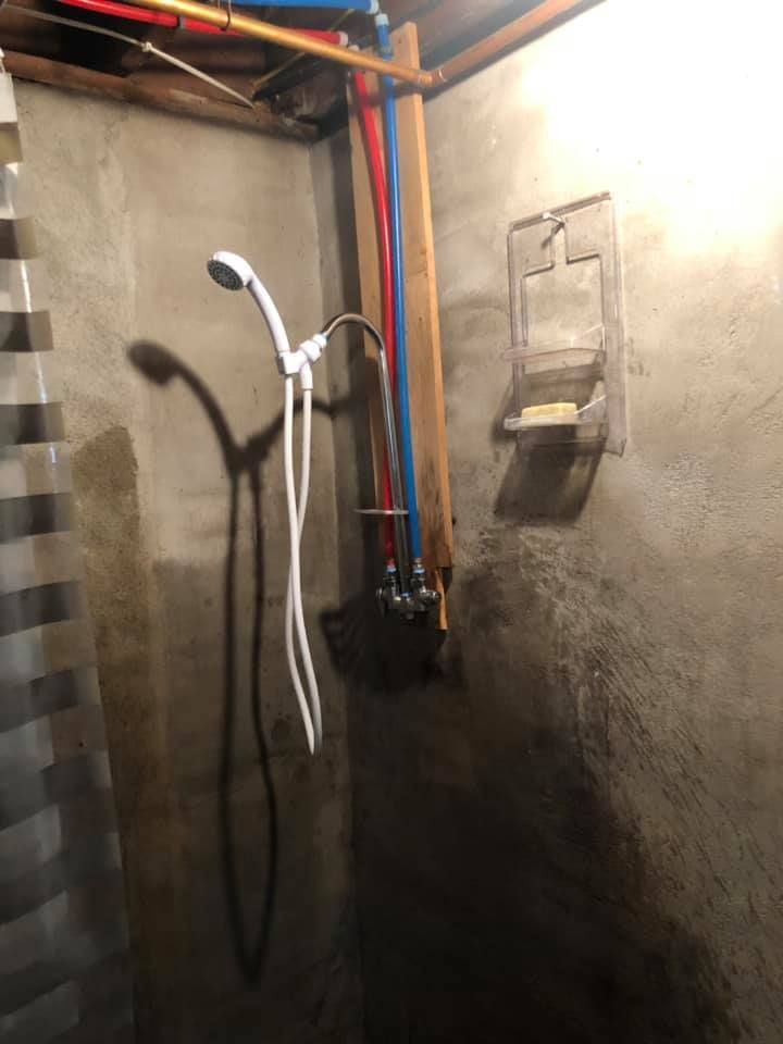Utility room shower setup with exposed pipes, a white shower hose, and a wall-mounted soap dish on concrete walls