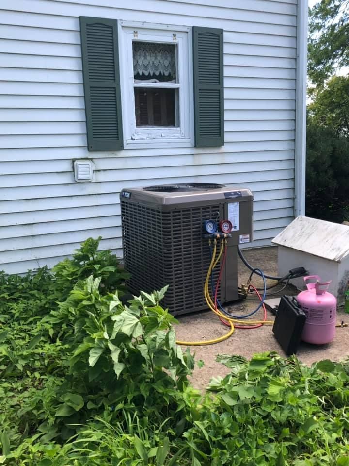 Outdoor HVAC unit beside a house wall with hoses and a pink refrigerant tank in overgrown grass