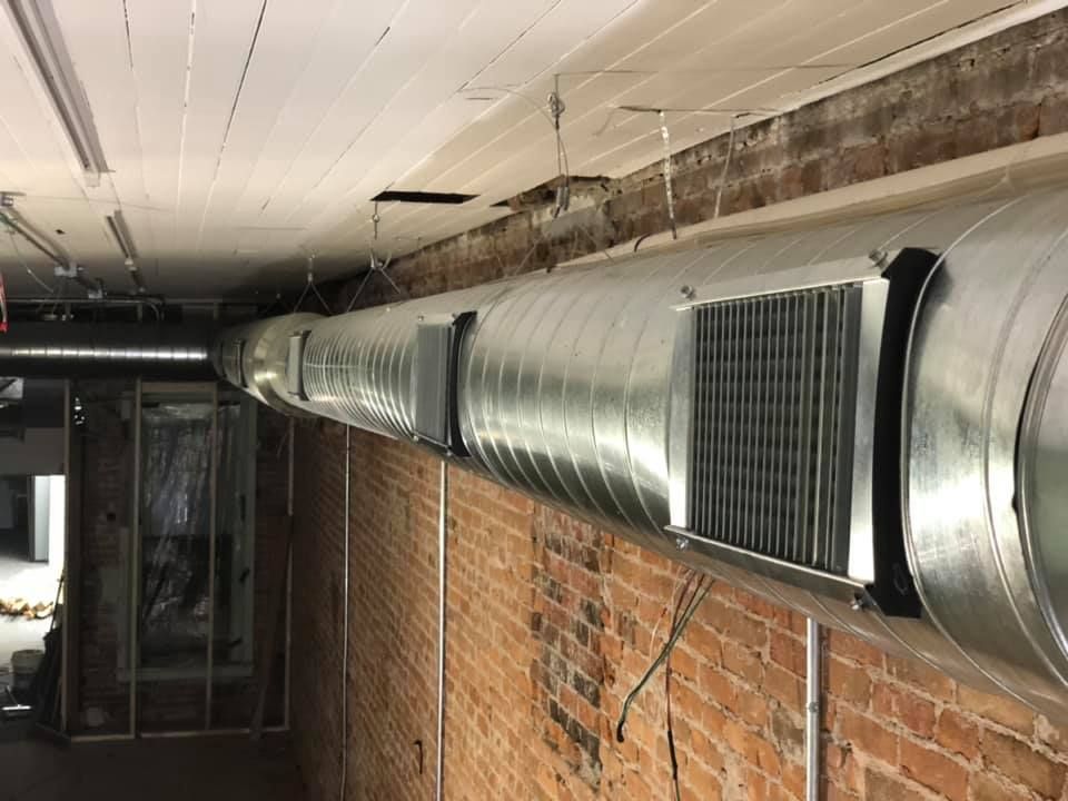 Long metal ductwork runs along a brick wall in an industrial basement corridor.