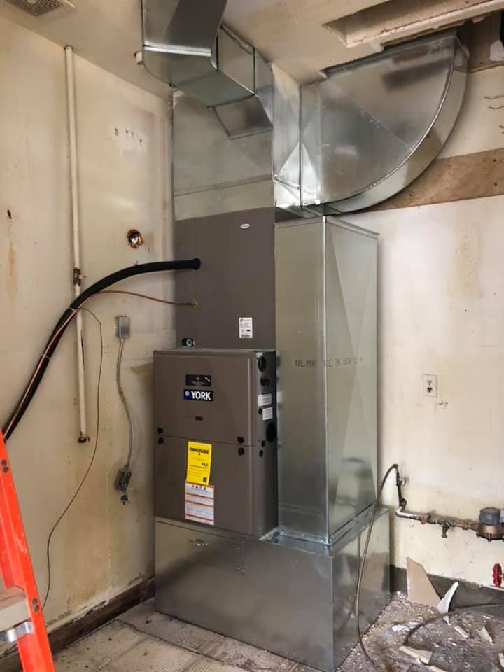 Gray HVAC/furnace unit with ductwork in a utility room corner, with exposed wiring and pipes.