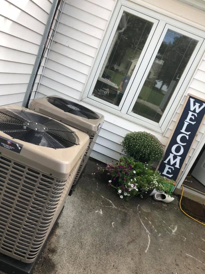 Two HVAC units beside a white house with a Welcome sign and potted flowers on the porch