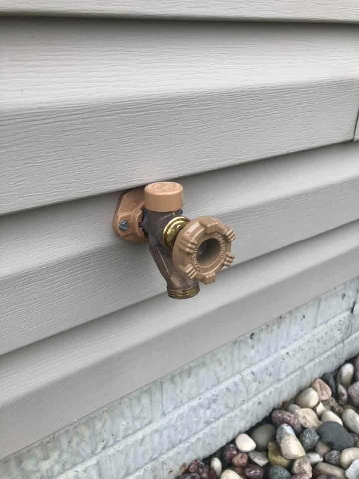 Outdoor brass hose spigot mounted on gray house siding above a stone foundation
