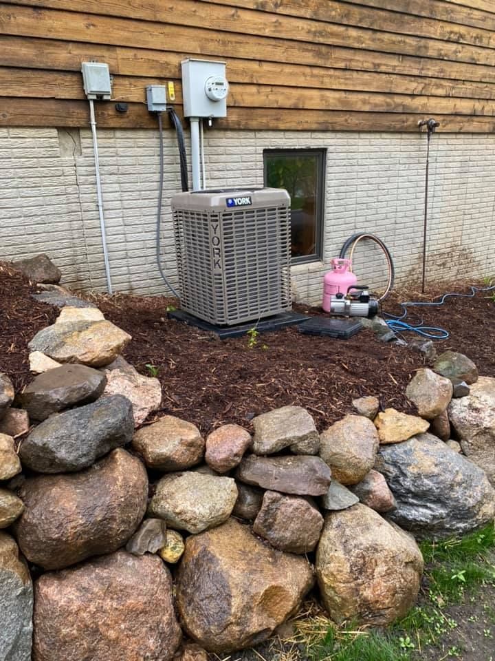 Outdoor HVAC unit beside a house wall, with utility meters and rocks in the foreground.