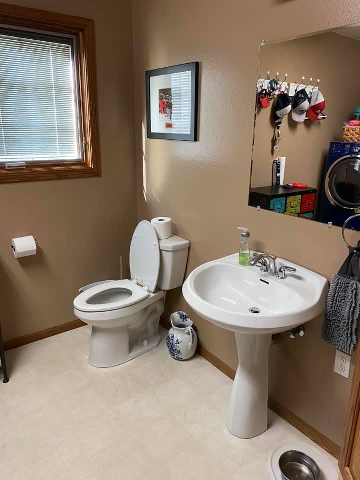 Small beige bathroom with toilet, pedestal sink, mirror, window, and towel hooks.