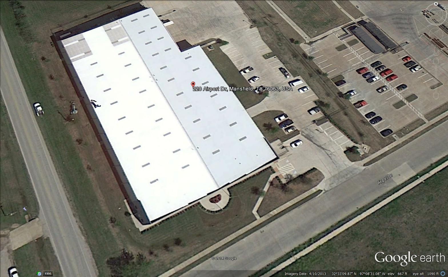 Aerial view of a large white-roofed building with parking lots and a road beside it.