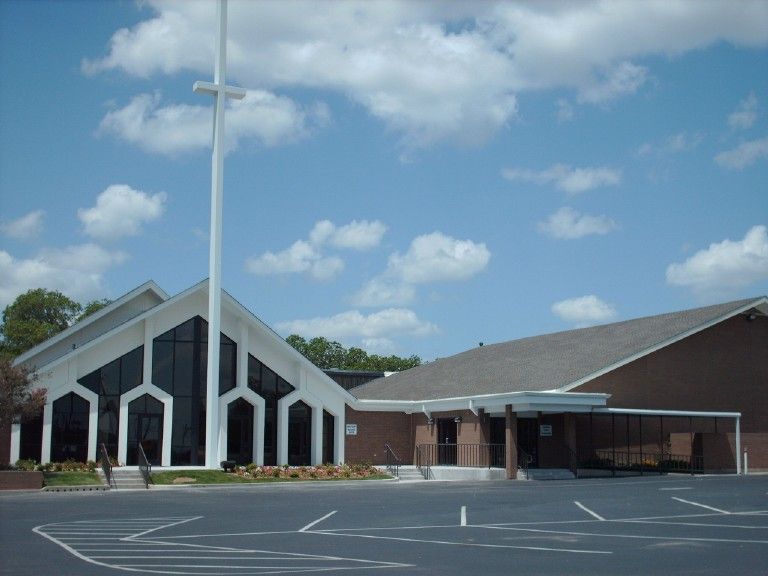 Church building with tall white cross, dark windows, and brick entrance under a blue sky