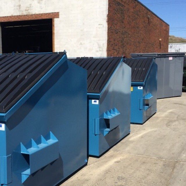 A row of blue dumpsters are lined up in front of a brick building