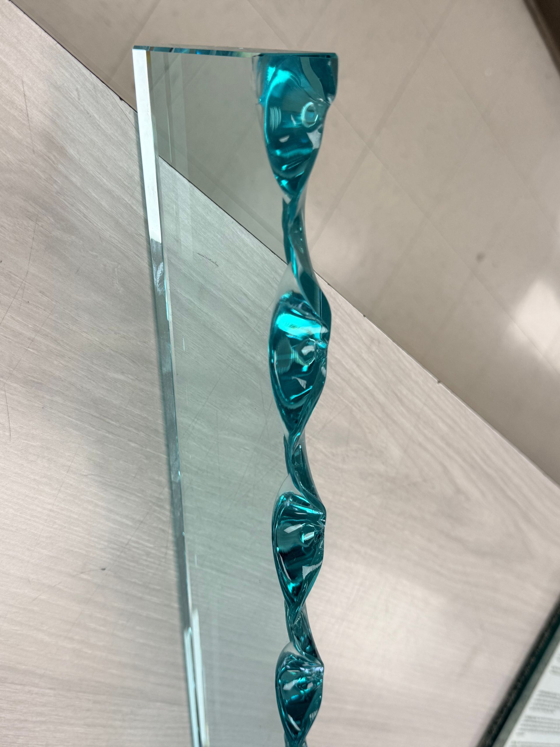 A clear glass award with a vertical, twisted teal blue core, standing on a light-colored surface.