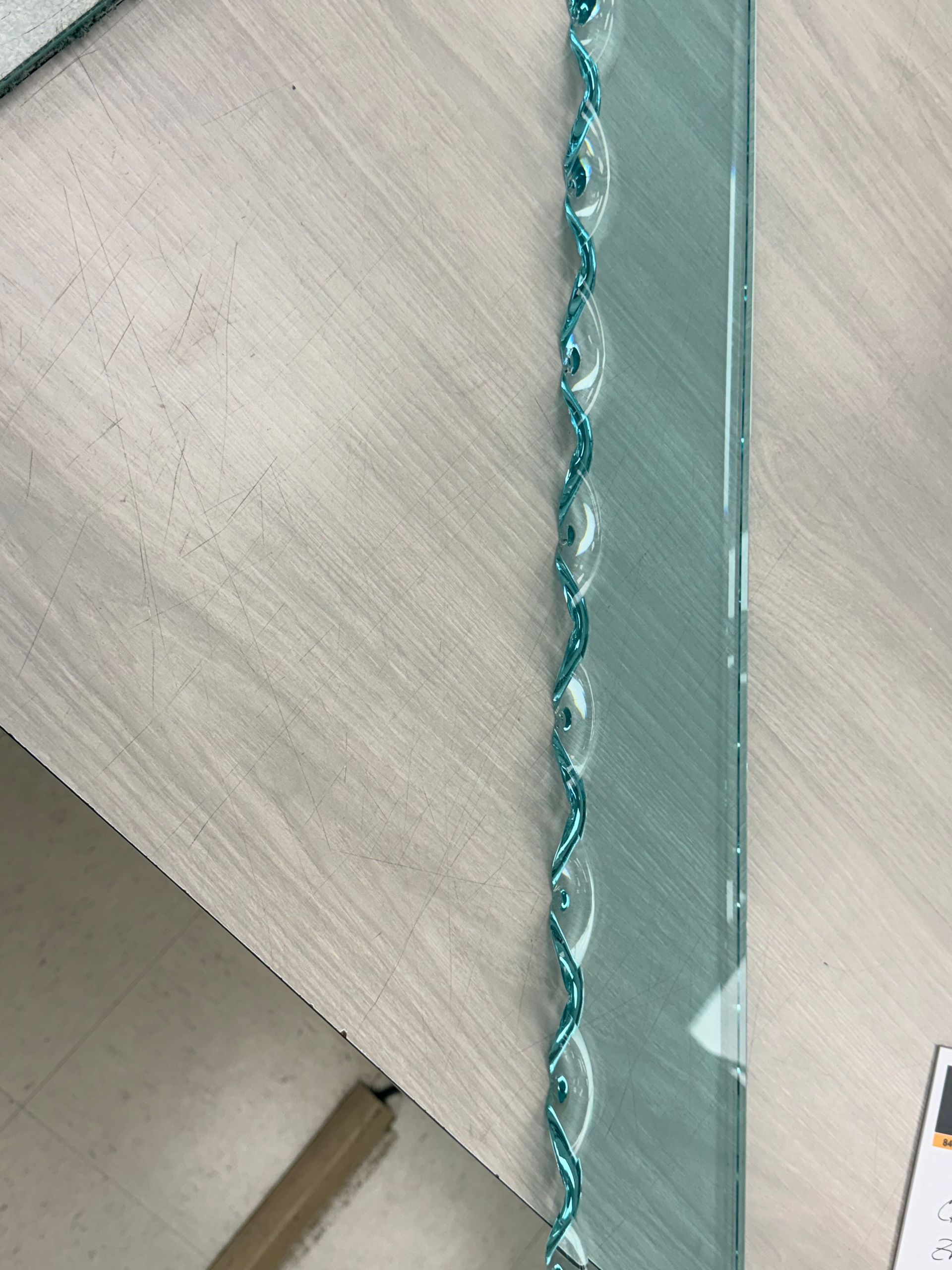 A rectangular piece of clear glass with a decorative, teal-colored braided or twisted edge design lying on a wood table.