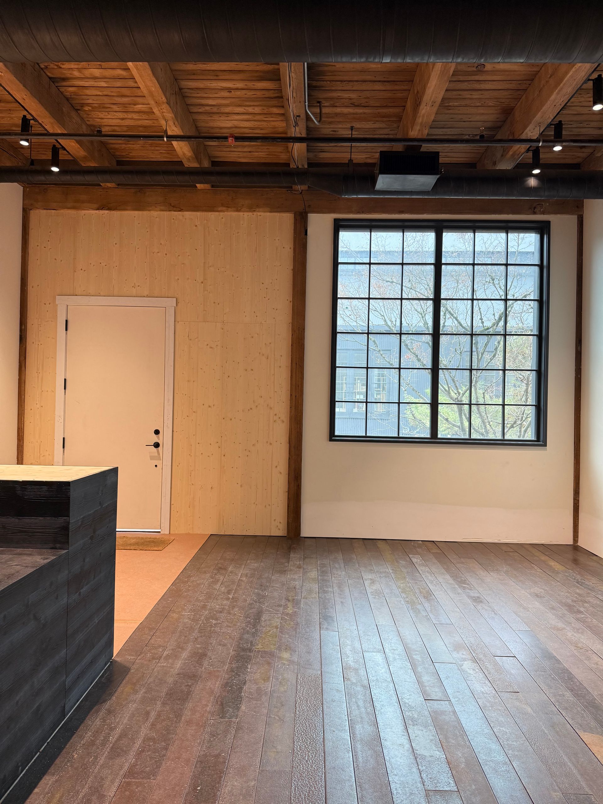 Empty room with wood floors, exposed ceiling beams, white walls, and a large window.