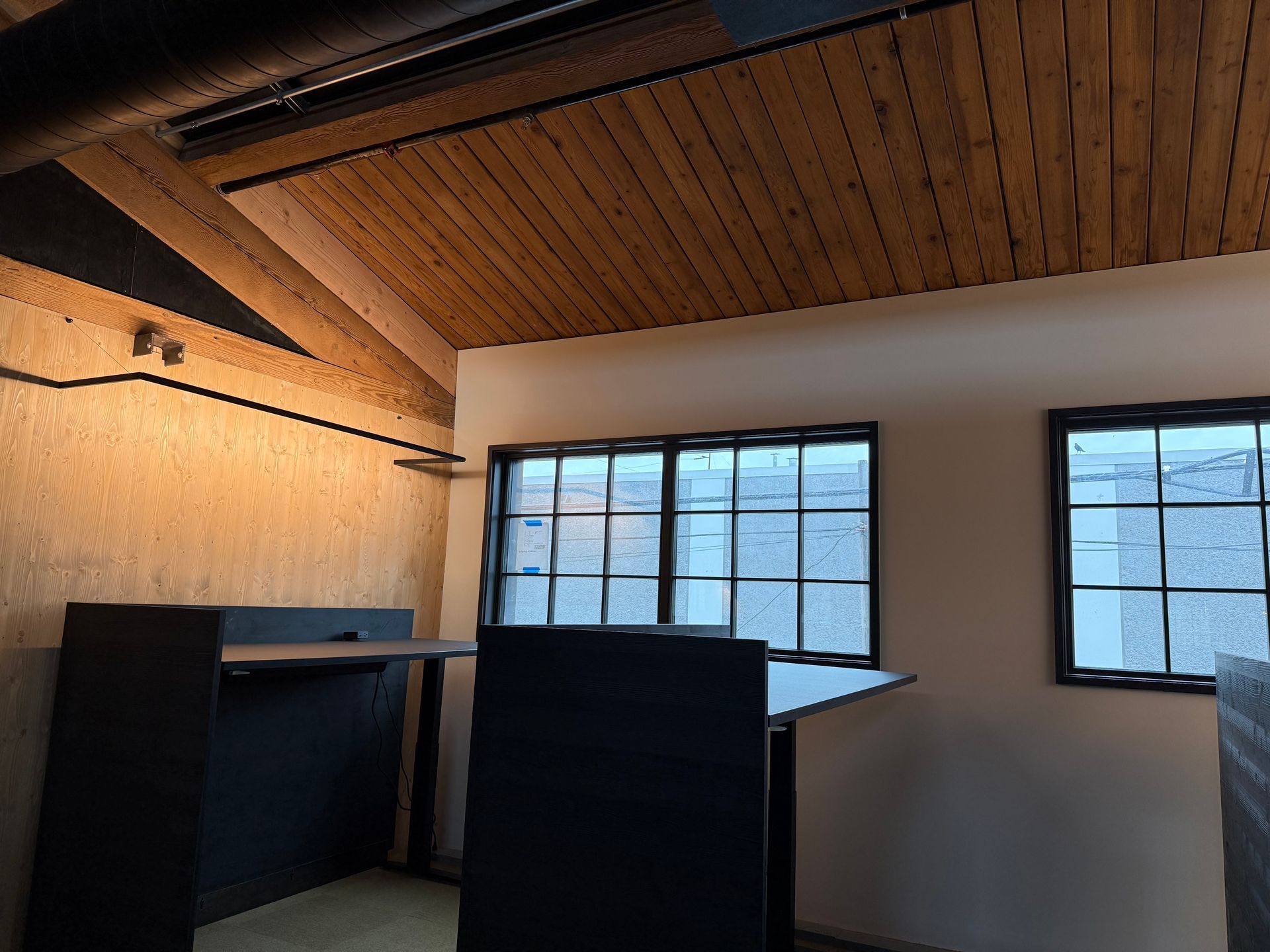 Empty room with wood-paneled ceiling, large windows, and dark cabinets or desks.