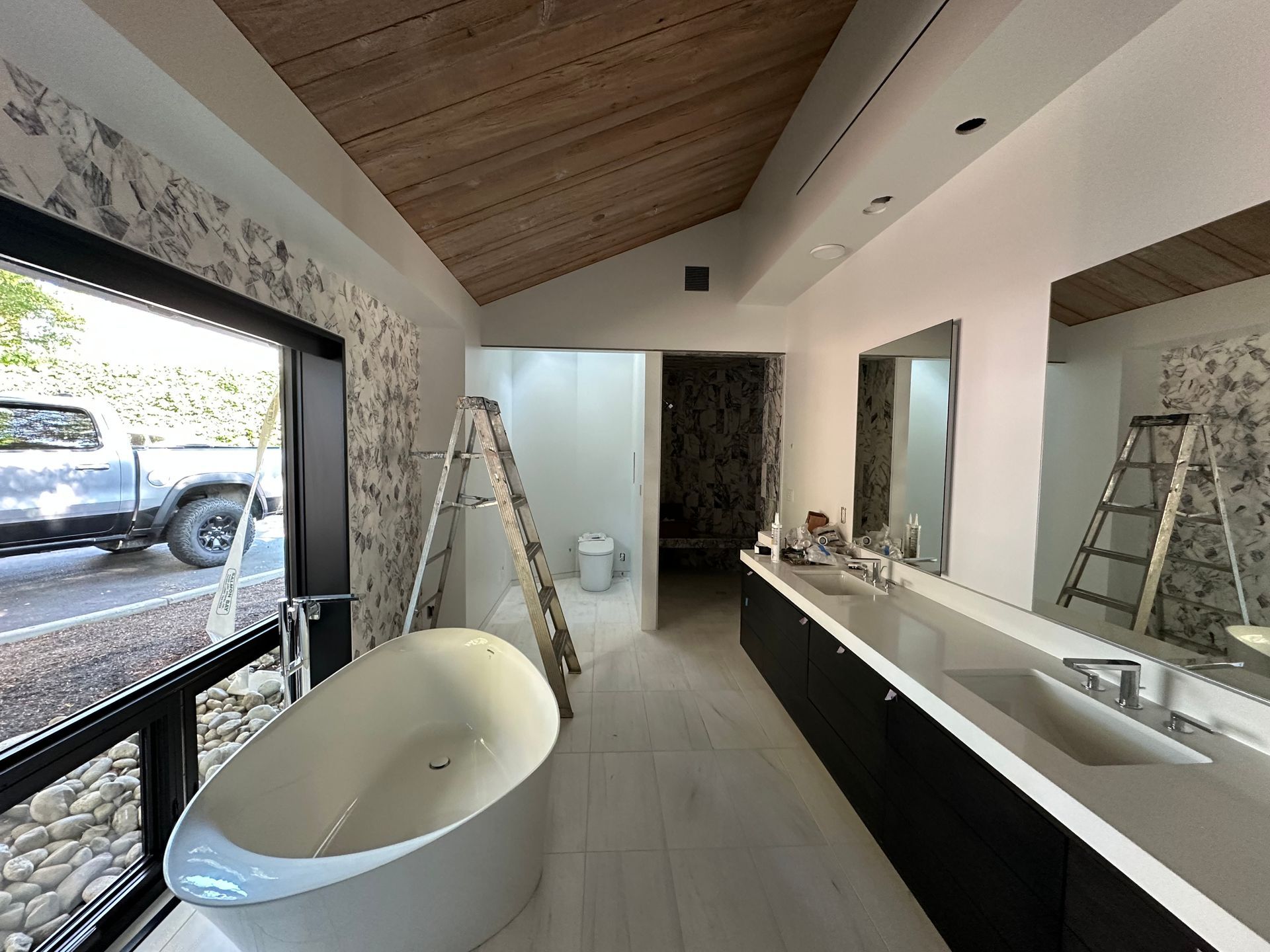 Modern bathroom under renovation with freestanding tub, long vanity, and ladders by large windows