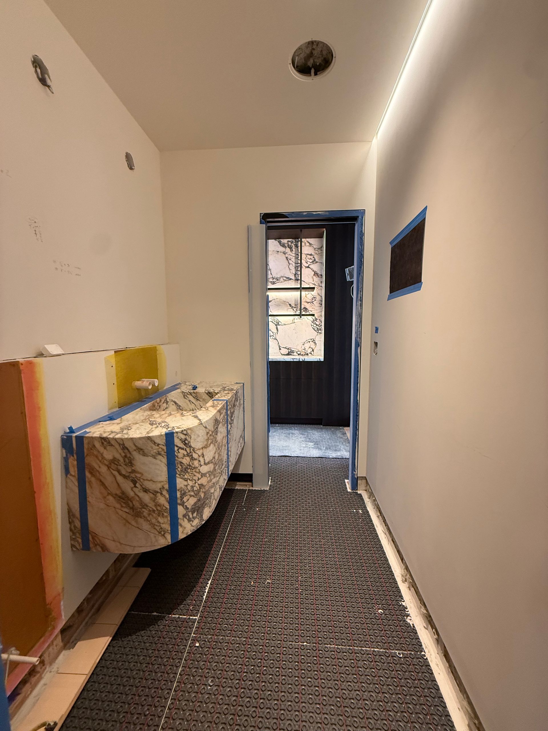 Narrow hallway with patterned floor covering, wrapped furniture on the left, and a doorway to a lit room ahead