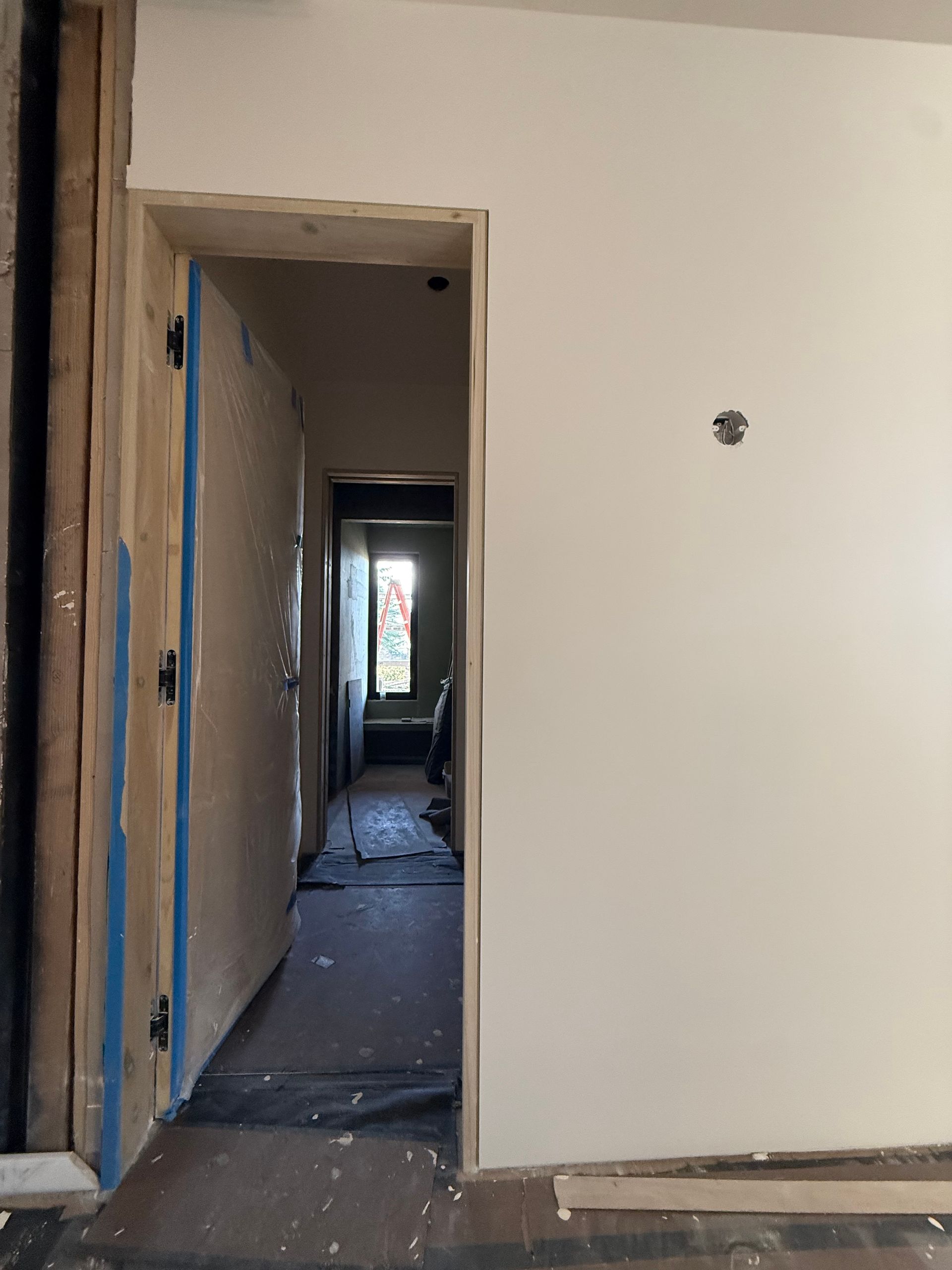Narrow hallway under renovation with unfinished walls, debris on the floor, and a doorway leading to a lit room.