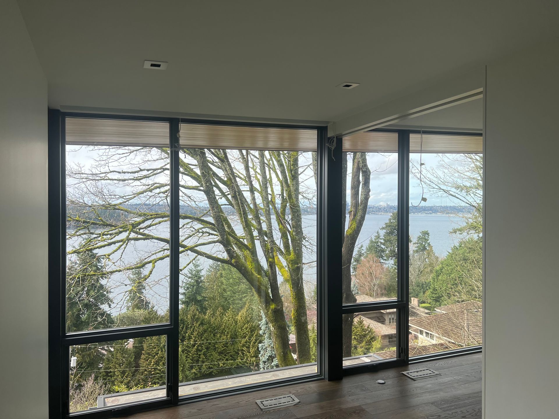 Large windows overlooking trees and a lake, with a white wall and wooden floor indoors