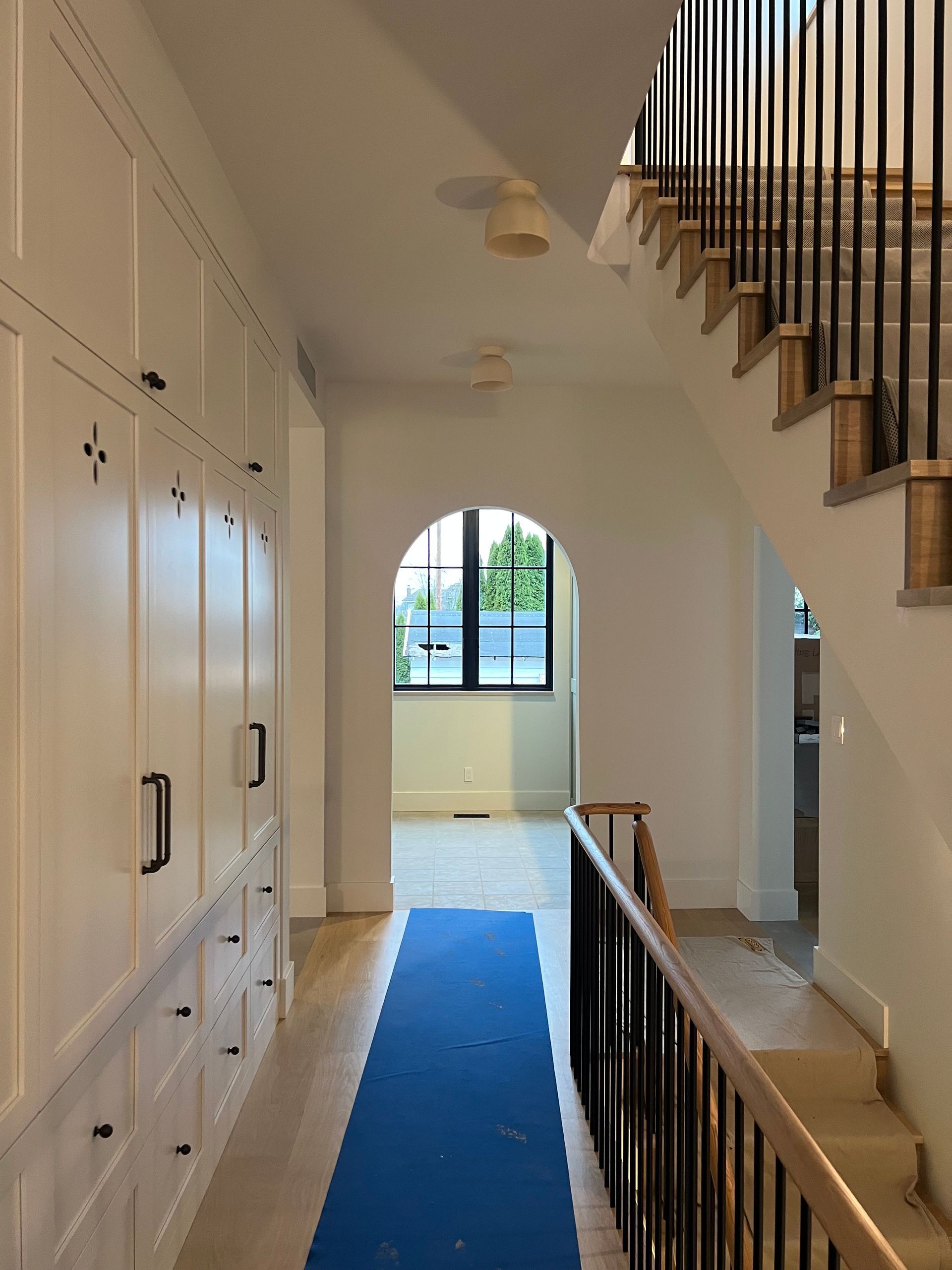 Bright hallway with blue runner, built-in cabinets, staircase railing, and arched window at the end.