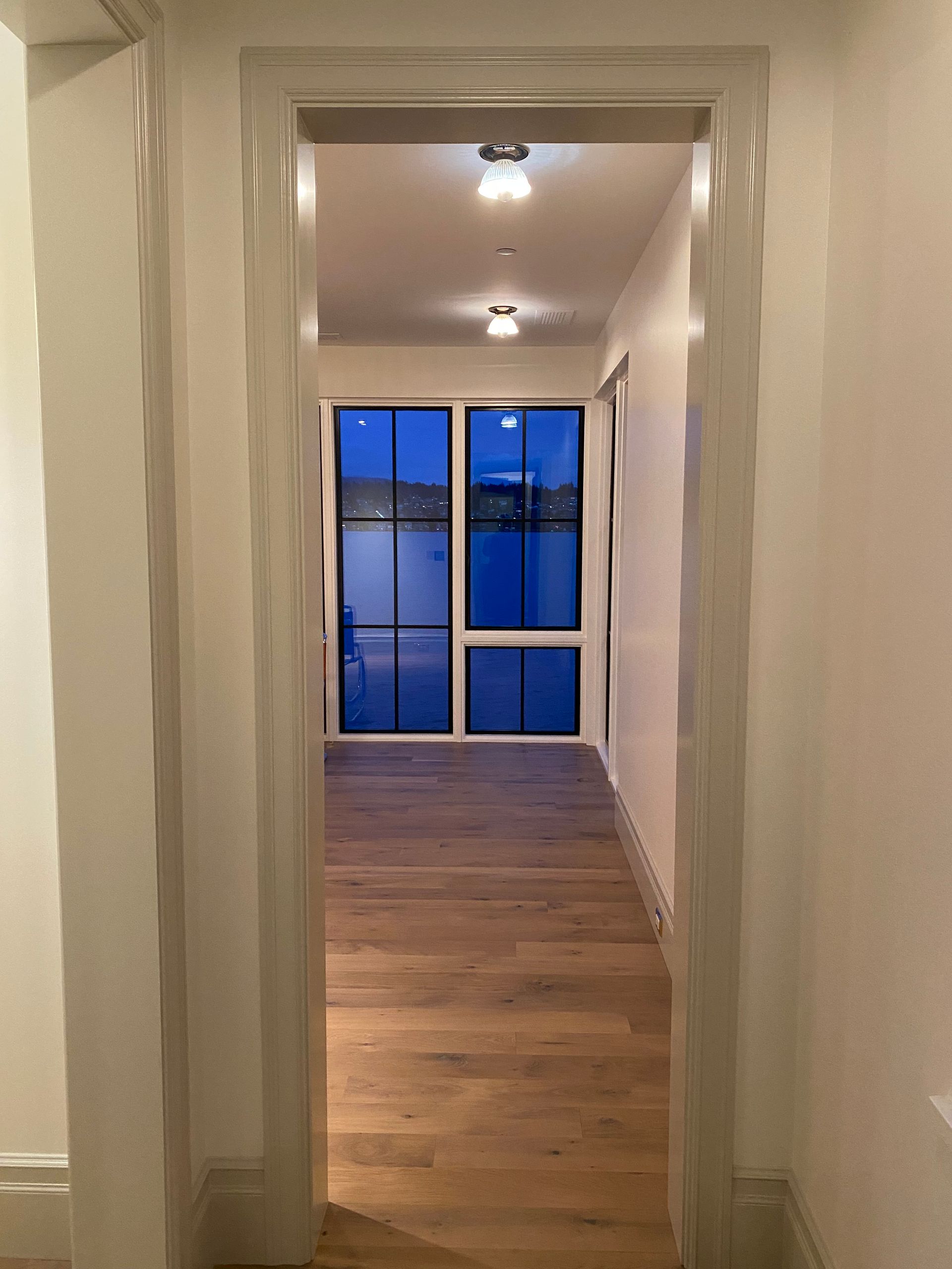 Hallway with wood floors leading to a glass door and windows lit by ceiling lights at dusk