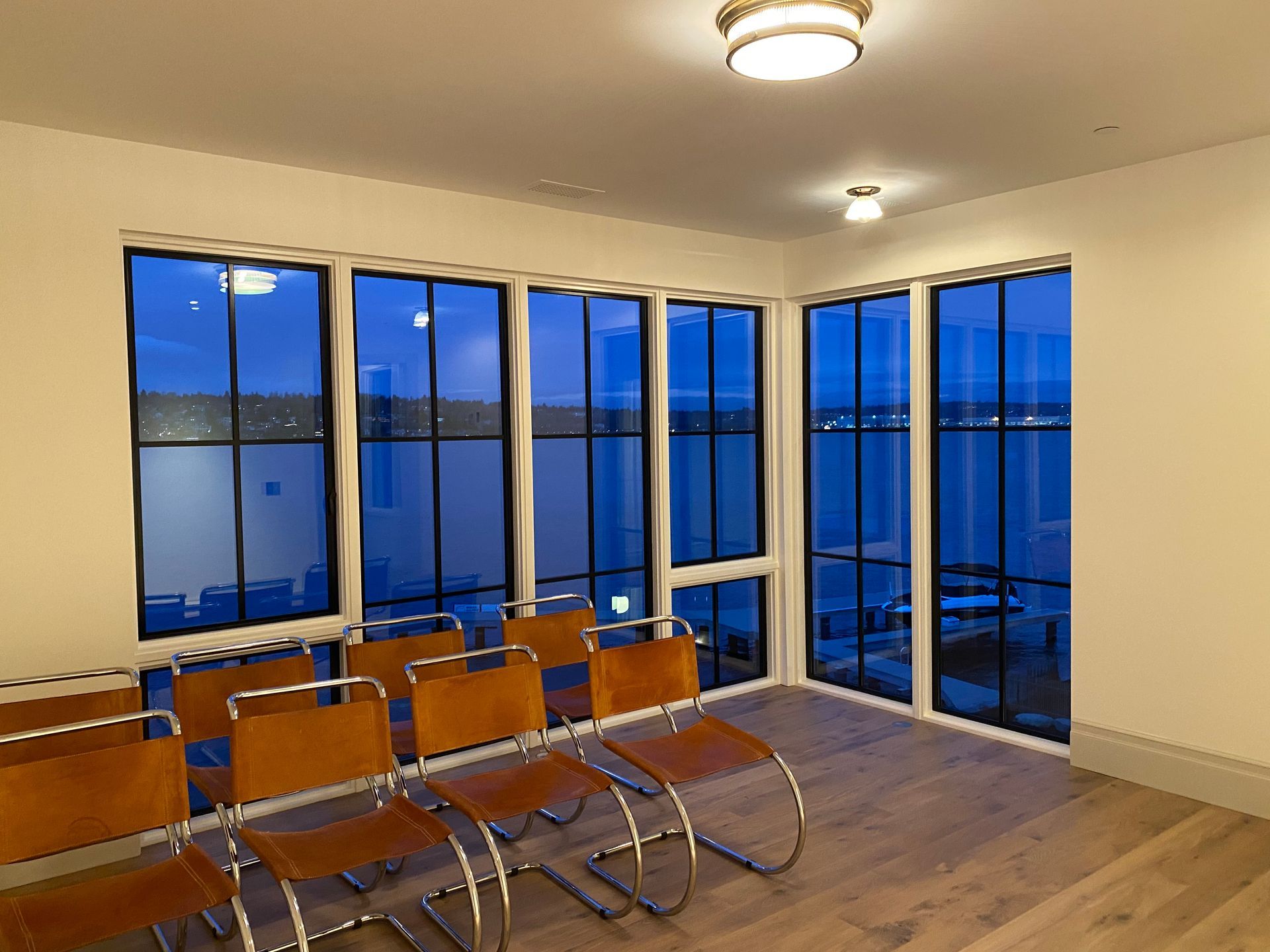 Empty modern room with chairs, wood floor, and floor-to-ceiling windows overlooking water at dusk