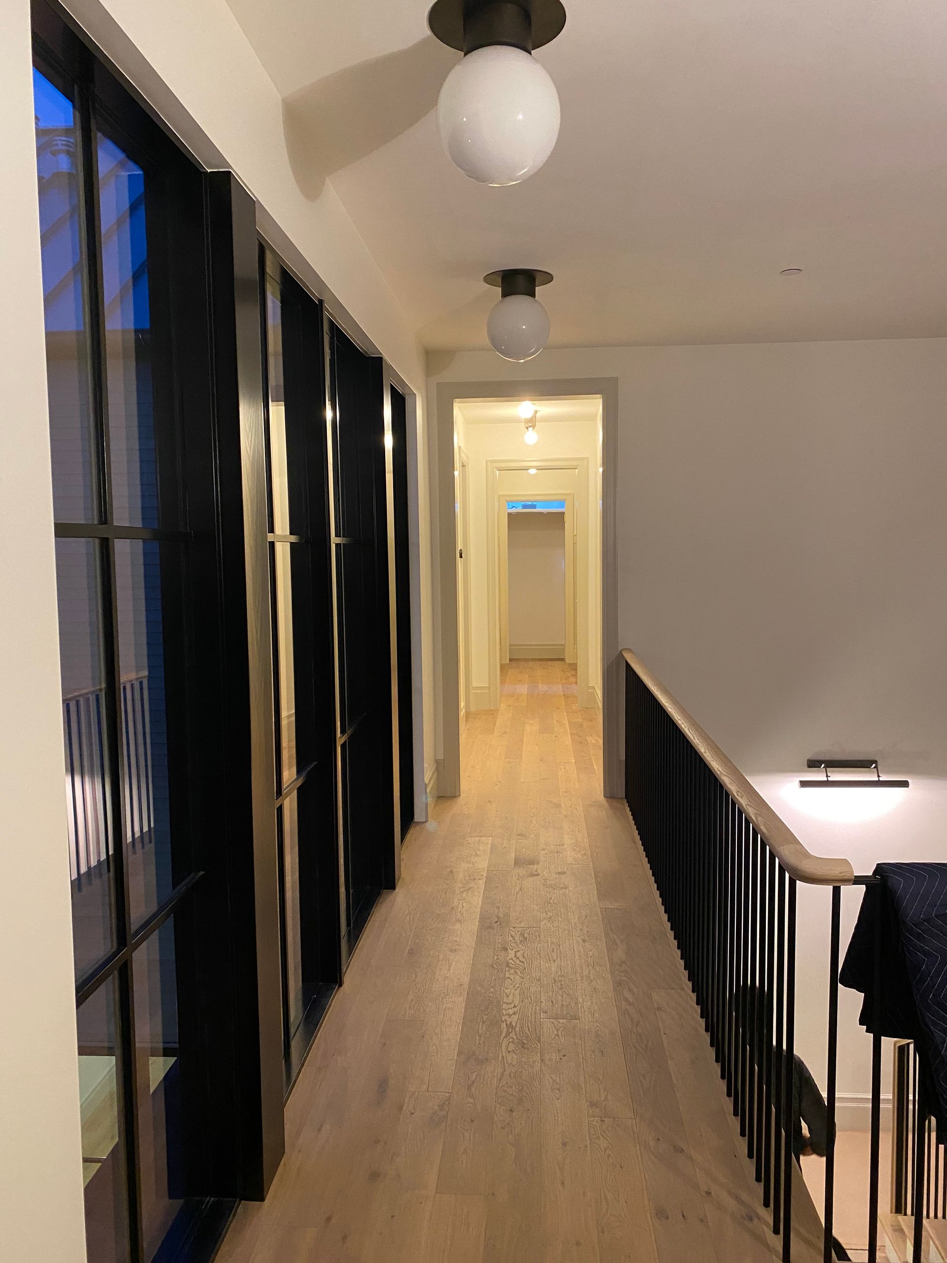 Narrow hallway with black-framed windows, hanging lights, and a railing overlooking a lower floor.