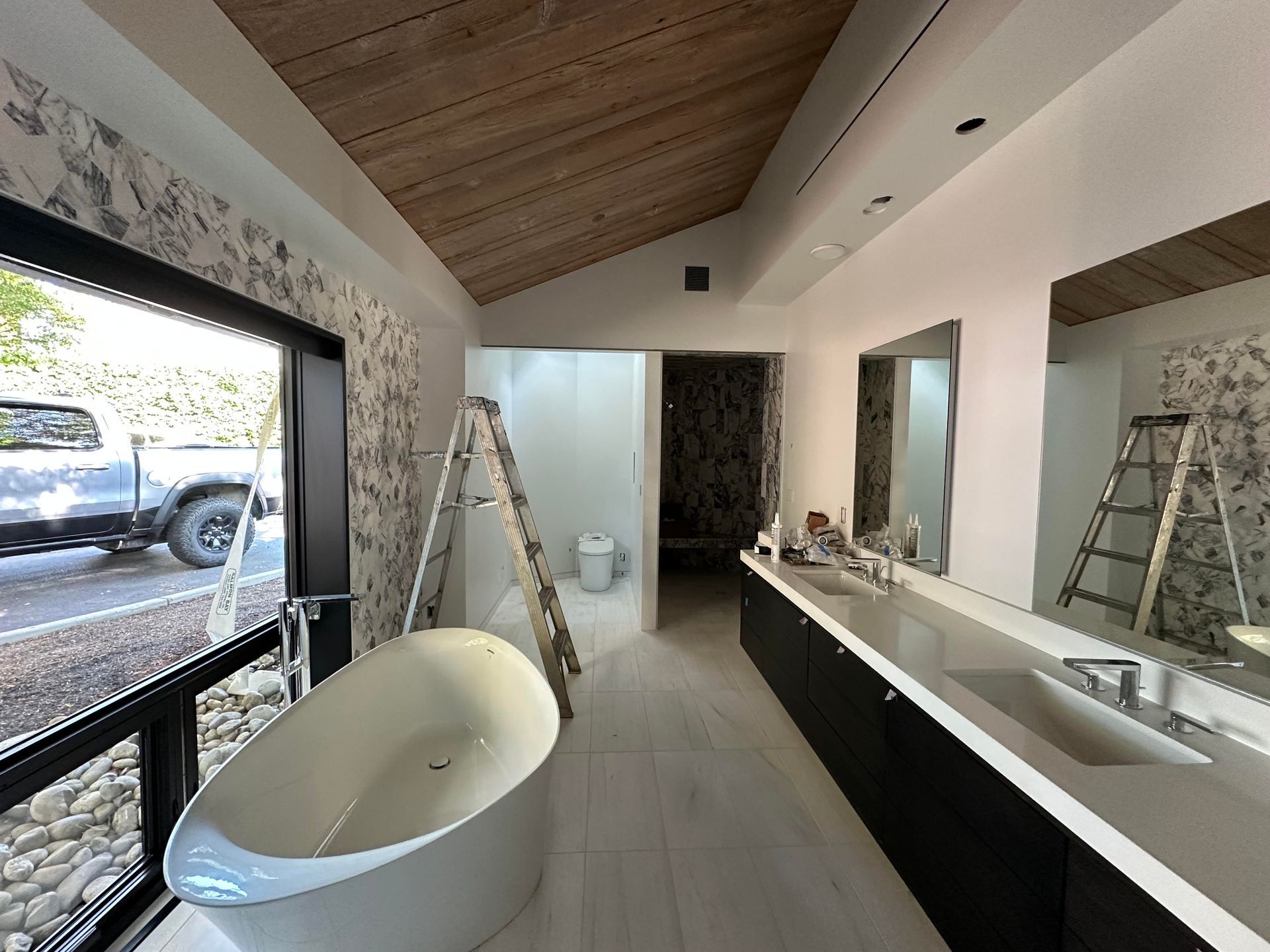 Modern bathroom under renovation with freestanding tub, long vanity, mirrors, and ladders near an open wall.