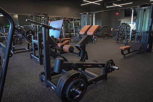 Empty gym with weight machines, benches, and free weights under dim overhead lighting.