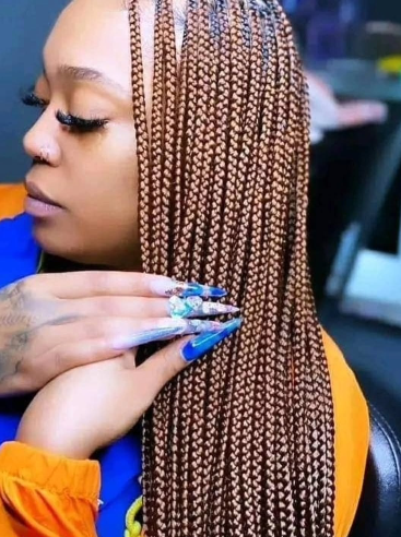 Woman with brown box braids, long nails with blue and white design, wearing orange and blue clothing.