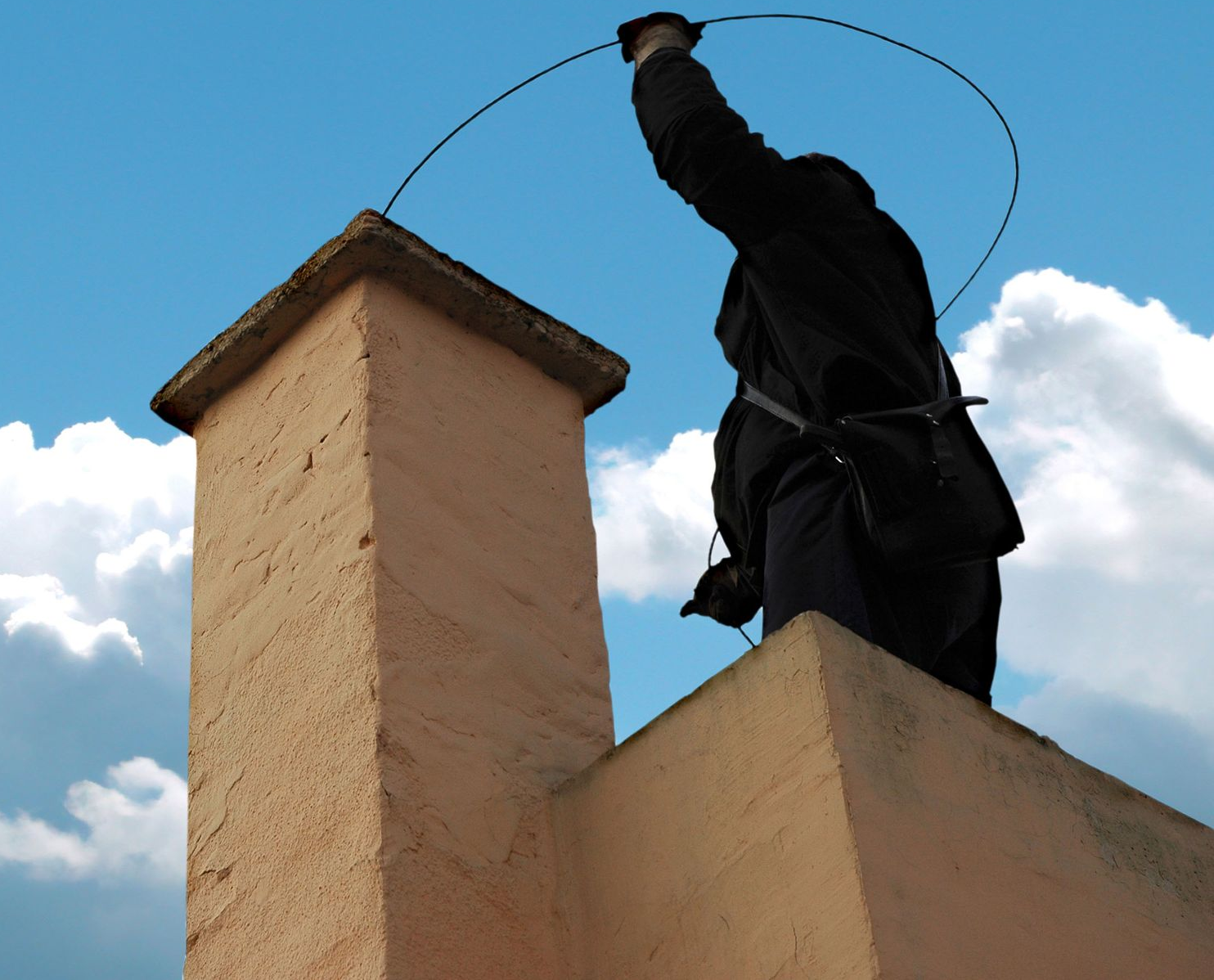 Person in black standing on a rooftop chimney, arm raised against a blue sky with clouds