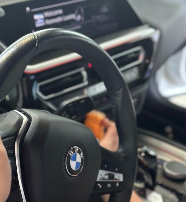 Black steering wheel with BMW logo, interior shot, person in car, infotainment screen visible.