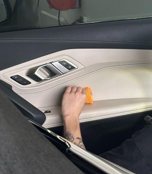 Person cleaning the beige interior door panel of a car with an orange sponge.